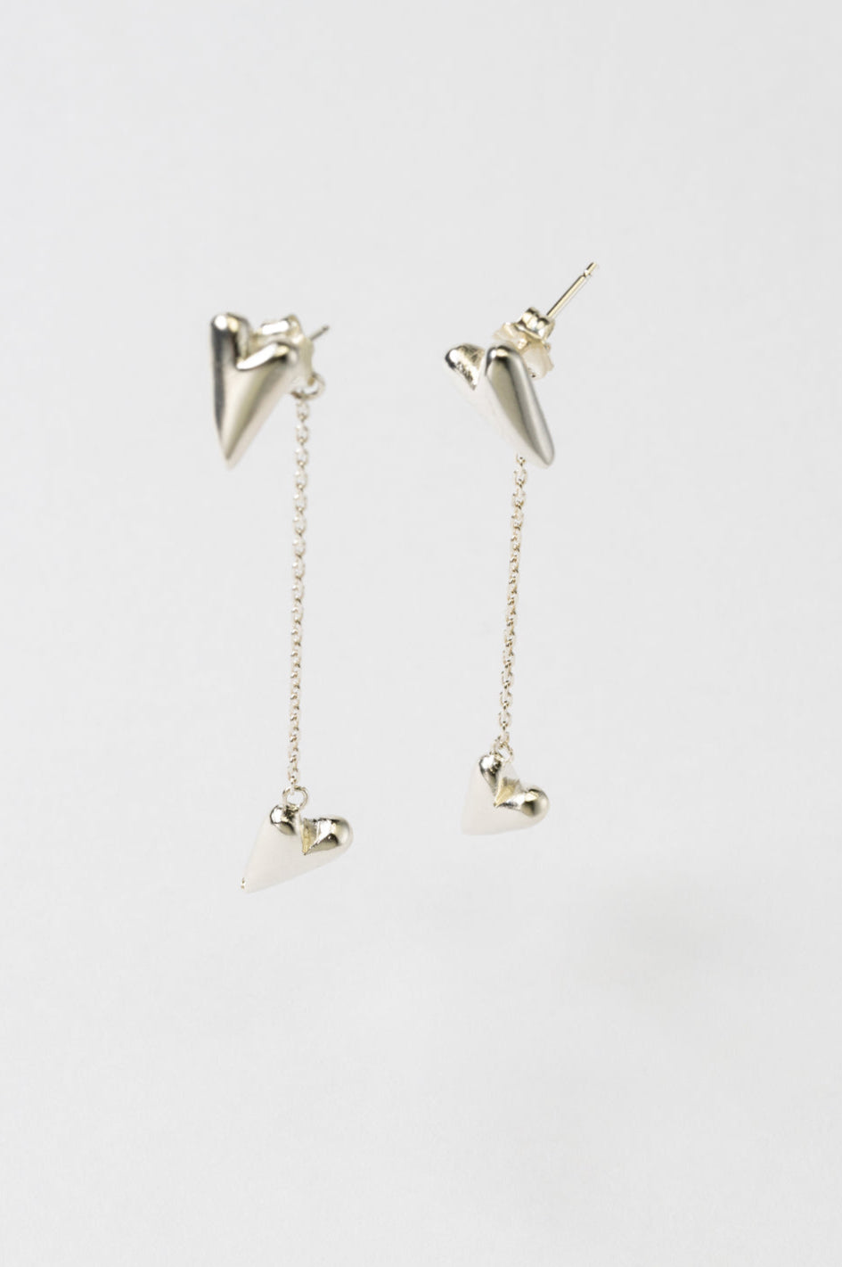 A pair of Silver earrings featuring a small heart on top connected by a chain to a longer heart-shaped pendant, displayed against a white background.